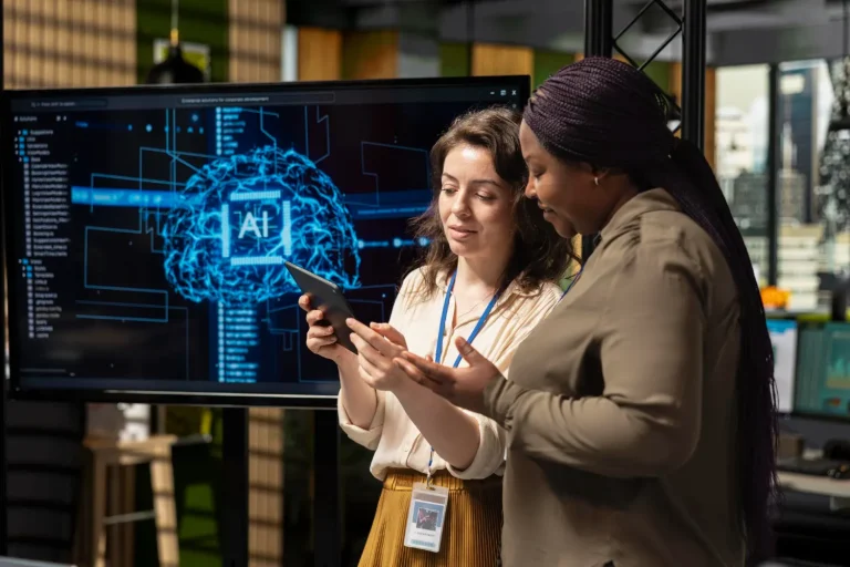 gemini para empresas: Duas profissionais em escritório analisam um aplicativo no celular enquanto uma tela com interface de IA e circuito digital aparece ao fundo, sugerindo inovação para empresas