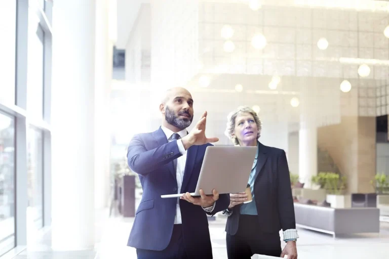 Homem de terno azul segurando notebook e explicando algo para mulher em ambiente corporativo moderno